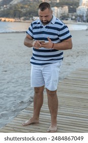 This image captures a man standing on a beach, engrossed in his smartphone. His casual attire suggests a leisurely day. 