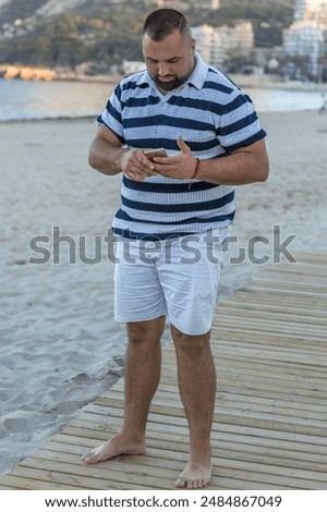 This image captures a man standing on a beach, engrossed in his smartphone. His casual attire suggests a leisurely day. 