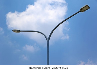This image captures a modern double-arm street light pole rising elegantly against a brilliant blue sky. Featuring a sleek, symmetrical curved steel design, it supports two LED luminaires extending in opposite directions. Backlit by a large, fluffy white cloud, this vital urban infrastructure ensures enhanced night visibility and safety for motorists. 