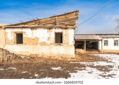 This image captures a mud-brick building with a partially collapsed wooden roof and broken windows. The house sits in a rural setting with patchy snow and dry ground highlighting the harsh environment