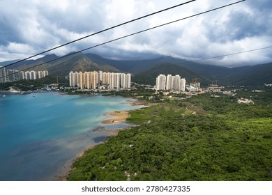This image captures the Ngong Ping 360 cable car system in Hong Kong, providing a panoramic view over Tung Chung Bay. A single cable car, possibly a Crystal Cabin with its distinct transparent features, is prominently suspended over the blue waters. In the background, the dense urban skyline of Tung Chung contrasts with the lush green hills of Lantau Island. The scene is set under a dramatic, cloudy sky, highlighting the 5.7-kilometer journey that connects downtown Tung Chung to Ngong Ping Village.