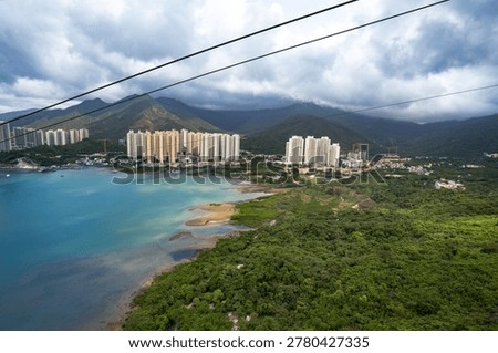 This image captures the Ngong Ping 360 cable car system in Hong Kong, providing a panoramic view over Tung Chung Bay. A single cable car, possibly a Crystal Cabin with its distinct transparent features, is prominently suspended over the blue waters. In the background, the dense urban skyline of Tung Chung contrasts with the lush green hills of Lantau Island. The scene is set under a dramatic, cloudy sky, highlighting the 5.7-kilometer journey that connects downtown Tung Chung to Ngong Ping Village.