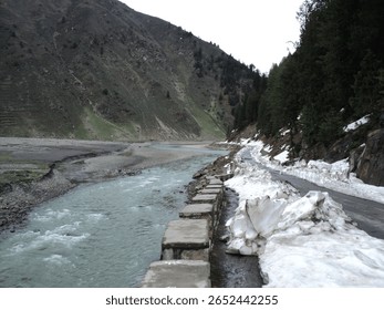 This image captures a powerful river flowing alongside a mountain road still partially obscured by significant snowdrifts, indicating the lingering effects of winter in a high-altitude valley 