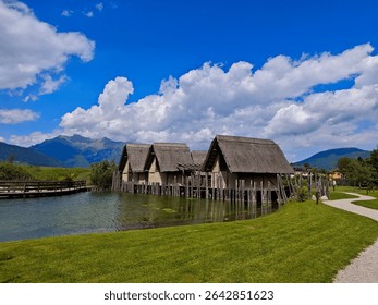 This image captures the reconstructed pile-dwelling village at Archeo Natura Park in Fiavé, Italy. Traditional thatched-roof houses stand on stilts over a tranquil lake, surrounded by lush green grass