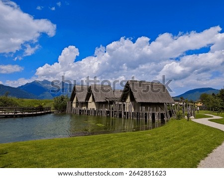 This image captures the reconstructed pile-dwelling village at Archeo Natura Park in Fiavé, Italy. Traditional thatched-roof houses stand on stilts over a tranquil lake, surrounded by lush green grass