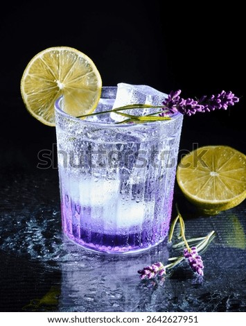 This image captures a refreshing and elegant lavender cocktail served in a textured glass with ice cubes. The drink shows a beautiful gradient from deep violet at the base .