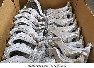 This image captures a scene inside an industrial workshop, showcasing numerous silver-white vehicle control arm metal castings neatly stacked inside a brown cardboard box. The components feature smooth and flat surfaces with a precise cast metallic texture, surrounded by protective bubble wrap. This reflects the standardized warehousing and pre-processing status of automotive parts before delivery, highlighting the mass production and quality control characteristics of automotive manufacturing.