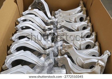 This image captures a scene inside an industrial workshop, showcasing numerous silver-white vehicle control arm metal castings neatly stacked inside a brown cardboard box. The components feature smooth and flat surfaces with a precise cast metallic texture, surrounded by protective bubble wrap. This reflects the standardized warehousing and pre-processing status of automotive parts before delivery, highlighting the mass production and quality control characteristics of automotive manufacturing.
