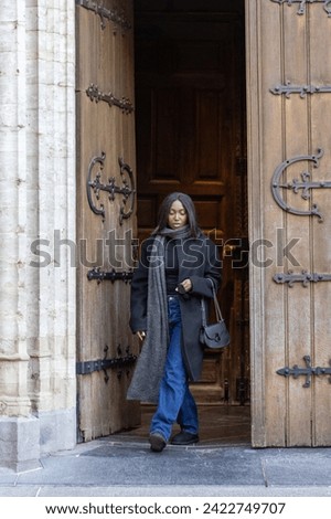 This image captures a serene moment as a young woman steps out from the dim interior of a gothic cathedral into the light. The heavy wooden doors adorned with intricate ironwork frame her, a symbolic