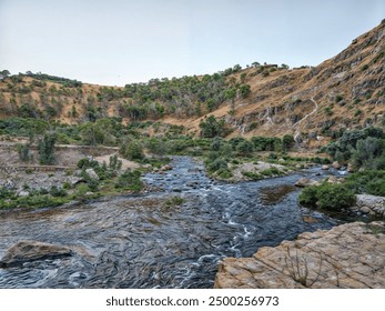  This image captures a serene natural scene featuring a small river flowing through a rocky mountainous area. The river is surrounded by lush greenery and rugged terrain. - Powered by Shutterstock