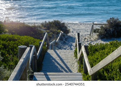 This image captures a serene wooden pathway leading down to a sandy beach, surrounded by lush green shrubs. The wooden handrails give a rustic feel, guiding the viewer’s eye towards the calm. - Powered by Shutterstock