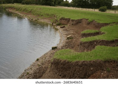 This image captures significant soil erosion along a lake bank revealing multiple layers of earth and green grass. The environmental degradation highlights the impact of natural forces.