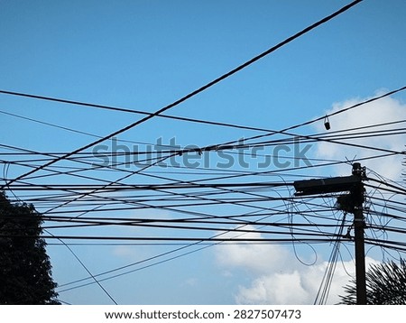 This image captures a strong visual contrast between nature and man-made infrastructure. In the foreground, a complex network of black electrical and telecommunications cables pierces the silhouette of a bright blue sky. The cables appear messy, tangled, and crisscrossed, creating a dense web of visual pollution.
On the right side of the image, an old wooden or concrete power pole appears at the center of all these connections. The pole appears worn, with the technical equipment atop it also appearing neglected, including a black junction box and tangled clumps of cables, adding to the clutter. Amidst the intricate web of cables, several small red dots are visible, possibly markers or connectors, as well as a small object hanging from the upper right-hand cable, likely part of an old piece of equipment or a leftover project.
