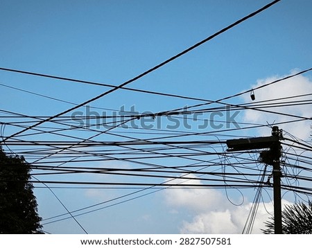 This image captures a strong visual contrast between nature and man-made infrastructure. In the foreground, a complex network of black electrical and telecommunications cables pierces the silhouette of a bright blue sky. The cables appear messy, tangled, and crisscrossed, creating a dense web of visual pollution.
​On the right side of the image, an old wooden or concrete power pole appears at the center of all these connections. The pole appears worn, with the technical equipment atop it also appearing neglected, including a black junction box and tangled clumps of cables, adding to the clutter. Amidst the intricate web of cables, several small red dots are visible, possibly markers or connectors, as well as a small object hanging from the upper right-hand cable, likely part of an old piece of equipment or a leftover project.