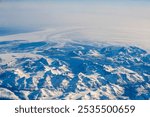 This image captures a stunning aerial view of snow-covered mountain peaks in Greenland, with a vast, icy landscape extending into the horizon.