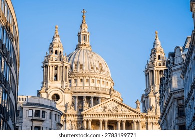 This image captures a stunning, low-angle view of St. Paul's Cathedral in London, bathed in the warm, golden light of either early morning or late afternoon. The iconic English Baroque dome dominates the center of the frame, flanked by its intricate western towers. The clear blue sky provides a vibrant backdrop that makes the weathered stone texture and the golden crosses atop the spires stand out beautifully. The cathedral is tightly framed between modern and classical city buildings on both sides, creating a compelling contrast between the historic monument and its urban surroundings.