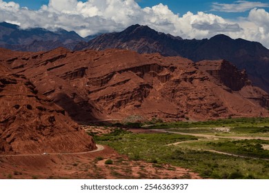 This image captures a stunning red rock canyon with dramatic formations, vibrant greenery, and a winding road beneath a backdrop of rugged mountains and clouds—perfect for nature and travel lovers. - Powered by Shutterstock