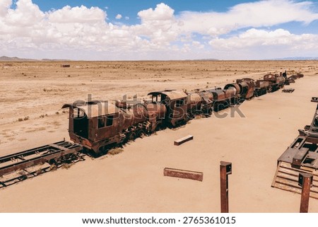 This image captures the surreal and desolate beauty of the Train Cemetery in Bolivia, a haunting reminder of a bygone era. A series of rusted and decaying locomotives and railcars stand abandoned in the arid desert landscape under a vast, expansive sky dotted with fluffy white clouds. The scene evokes a sense of decay and the passage of time, as nature reclaims these once-powerful machines. The harsh sunlight casts long shadows, accentuating the textures of the rusted metal and the dry, cracked earth. The composition highlights the contrast between the industrial remnants and the natural environment, creating a visually striking and thought-provoking image. The setting showcases the stark beauty and isolation found in the heart of Bolivia, where the echoes of history linger in the wind. The train graveyard serves as a unique travel destination and a testament to the challenges and triumphs of early transportation in a remote and unforgiving landscape. It stands as a poignant symbol of progress and the relentless march of time, inviting reflection on the industrial past amidst the stunning scenery of Bolivia. The image is rich in texture and detail, inviting viewers to explore the layers of history and the stark beauty of the arid environment in Bolivia.