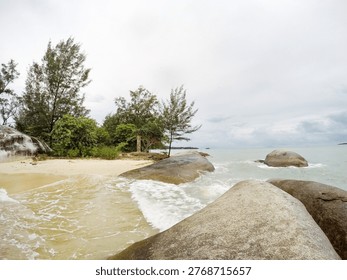 This image captures a tranquil beach scene with large rocks and trees on a cloudy day. The location appears to be a secluded cove with a mix of sandy shore and rocky coastline. The ocean waves gently lap against the rocks, creating a soothing melody. The cloudy sky adds to the serene atmosphere, making it an ideal spot for relaxation and contemplation. The lush greenery and trees in the background provide a sense of seclusion and privacy, while the vast expanse of the ocean stretches out to the horizon.