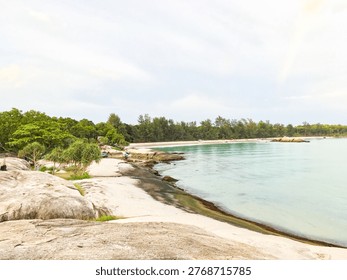 This image captures a tranquil beach scene with large rocks and lush greenery along the shoreline. The calm turquoise waters stretch out to the horizon, meeting a cloudy sky. The serene atmosphere makes it an idyllic destination for relaxation and recreation. Located in a tropical paradise, this beach offers a picturesque getaway from the hustle and bustle of daily life.