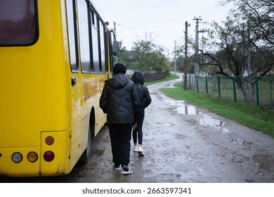 This image captures two people walking toward a bright yellow bus parked on a muddy road after rainfall. The scene shows a rural or suburban area with a mix of nature and infrastructure.