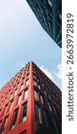 This image captures an upward perspective of modern architecture,showcasing the sharp corner of a tall red building with a grid of rectangular windows.The sunlight casts clean shadows on its geometric