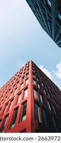This image captures an upward perspective of modern architecture,showcasing the sharp corner of a tall red building with a grid of rectangular windows.The sunlight casts clean shadows on its geometric