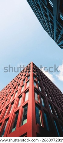 This image captures an upward perspective of modern architecture,showcasing the sharp corner of a tall red building with a grid of rectangular windows.The sunlight casts clean shadows on its geometric