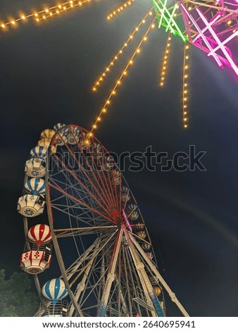 This image captures a vibrant night scene at what appears to be a carnival or fair. The main subject is a large Ferris wheel, brightly lit with red and blue lights, contrasting against the dark night 
