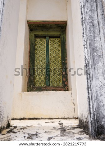 This image captures the weathered stone and ornate gold filigree of a traditional Thai temple window, offering a view into the country's rich architectural and spiritual history. The vibrant green and gold patterns stand out against the aged white walls, blending decay with enduring beauty.