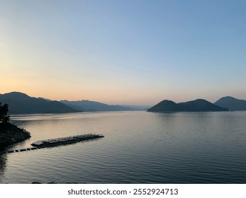 This image captures a wide, tranquil lake during either sunset or sunrise, surrounded by distant mountains. The horizon is bathed in soft, warm tones from the fading or emerging sunlight.