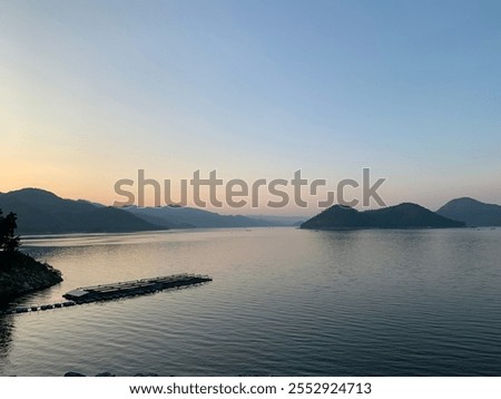 This image captures a wide, tranquil lake during either sunset or sunrise, surrounded by distant mountains. The horizon is bathed in soft, warm tones from the fading or emerging sunlight.
