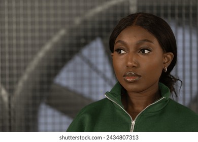 This image captures a young African American woman in a green zip-up jacket, poised against a blurred geometric patterned background. Her expression is pensive, her gaze directed away from the camera - Powered by Shutterstock