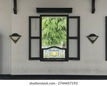 This image centers on a single open window set within a clean white wall, framed by dark wooden shutters that extend outward on both sides. The composition is symmetrical, with matching decorative wall sconces positioned evenly to the left and right. The lower portion of the window features a stained-glass panel with intricate geometric and floral motifs in soft blues, yellows, and greens, adding a delicate ornamental accent. Through the opening, lush green foliage is visible outside, creating a vivid contrast against the neutral interior tones. The scene feels calm and balanced, highlighting architectural detail, craftsmanship, and the interplay between interior space and the natural world beyond.