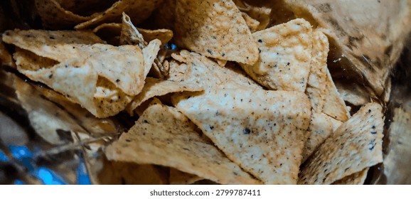 This image is a close-up of a pile of triangular tortilla chips, like nacho chips. They’re golden-orange with a slightly rough, bumpy texture and scattered dark specks of seasoning, maybe chili or pepper. Some chips overlap, while others are broken or bent at the edges, showing how crispy they are. 

The lighting is warm and a bit dramatic, casting soft shadows that highlight the ridges and texture on each chip. The background is dark and out of focus, with a hint of blue and the crinkled edge of a snack bag, so all attention stays on the chips. 

The overall feel is crunchy and savory, like a snack ready to dip into salsa or guac.
