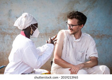 In This Image, A Content White Caucasian Healthcare Worker Is Receving A Vaccine Shot By A Black Female Doctor During Infectious Disease Prevention In Africa