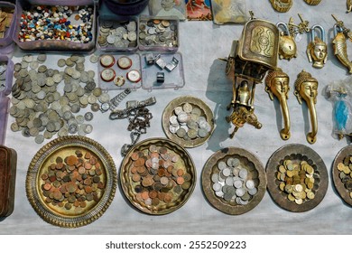 This image depicts a diverse array of items such as coins, jewelry, and brass artifacts laid out on a table. The items include old coins, watches, and decorative handles, indicating a flea market or a