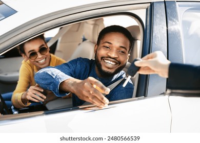 This image depicts a happy couple receiving the keys to their new car from a dealership representative. The man is sitting in the drivers seat of a white sedan and is reaching out to take the keys