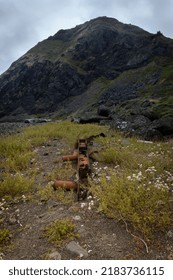 This Image Depicts An Old Rusty Car Part Pointing Towards A Large Mountain. The Foreground Has A Warm Lighting Cast Upon It, But The Mountain Has Cold Lighting That Further Dramatizes Its Size.