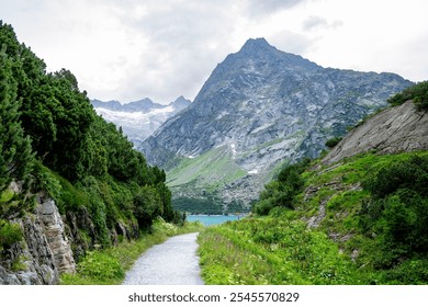 This image depicts a scenic mountain path in Switzerland, leading toward a valley with lush greenery and a glimpse of a turquoise lake. Towering rocky peaks, partially covered with patches of snow. - Powered by Shutterstock