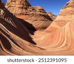 This image depicts stunning sandstone formations with smooth, wave-like patterns in a desert landscape under a bright blue sky. The red and orange hues create a mesmerizing geological spectacle.