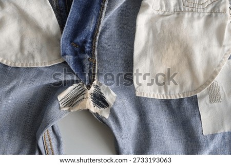 This image displays the inside out view of a pair of light blue denim jeans, highlighting a repair near the crotch area. The repair at the crotch of the jeans involves a small, irregularly shaped patch of white or off-white fabric applied to the inside of the garment. The patch has been secured using a running stitch, which are the rows of thick, dark-colored parallel lines visible on the fabric. The technique used, involving a basic stitched patch on the reverse side of the worn area, is a common and durable method for mending high-stress areas to prevent further tearing.