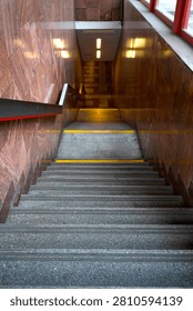 This image is a dynamic, high-angle perspective looking directly down a long flight of concrete stairs descending deep into a public underground passageway, likely a metro station or building entrance. The concrete steps are textured, showing wear and safety treads, with bright yellow hazard markings on several levels for safety. The stairwell is defined by polished, reflective walls clad in distinctive reddish-brown and pinkish-red natural stone or marble panels, creating a warm, deep color scheme and reflecting the light. A prominent, dark-painted metal handrail with a thin red accent strip runs along the left side. Several rectangular, warm fluorescent light fixtures are visible further down, illuminating the tunnel-like passage below and creating a sense of distance and destination. A window with a bright red frame is visible at the very top right, contrasting with the interior's warmth. The overall composition is leading lines, drawing the eye vertically into the shadowy depth, creating a feeling of descent, urban exploration, and movement. The natural light from above mixes with the artifical light below. This photograph captures the essence of urban transportation infrastructure, interior architecture, and everyday transit experiences, emphasizing form, texture, and a unique atmosphere.