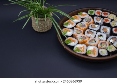 This image exudes a sense of Zen with its simple yet stylish presentation of a variety of sushi rolls in a circular wooden tray, positioned next to a potted green plant in a woven basket. The dark bac - Powered by Shutterstock