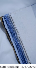 This image features a closeup, angled shot of a handmade journal or sketchbook, highlighting its unique textures and craftsmanship.

The most striking feature is the thick, deckle-edged paper. The edges are rough and uneven, characteristic of traditional hand-molding processes. The pages transition in color from a crisp off white on top to various shades of cornflower and cobalt blue deeper in the stack.