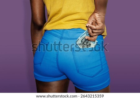 This image features a close-up, back-facing view of a person wearing vibrant blue, tight-fitting shorts and a yellow shirt against a solid purple background. The person’s right hand is reaching into their back pocket, pulling out or stashing a bundle of Ghanaian Cedi (GHS) banknotes. The teal and blue hues of the notes, typically associated with higher denominations like the 100 or 200 cedi bills, are visible. The composition highlights themes of West African finance, personal wealth, and a bold, colorful aesthetic common in contemporary urban photography.