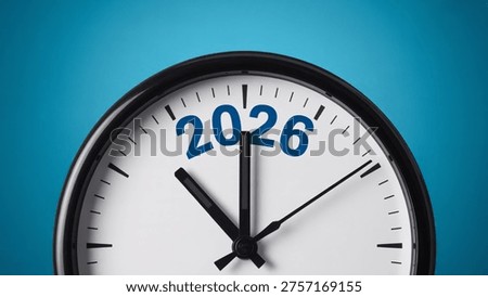 This image features a close-up of a clock face with the year 2026 displayed prominently on a blue background. The clock has a sleek and modern design with a black frame and white face. The hour and minute hands are black, and the tick marks are thin and evenly spaced. The image conveys a sense of time, schedule, and planning, and could be used to represent concepts such as productivity, efficiency, and goal-setting.