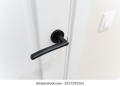 This image features a closeup view of a sleek black door handle that is mounted on a pristine white door, highlighting the contrast between the two colors and the simple elegance of the fixture - Powered by Shutterstock