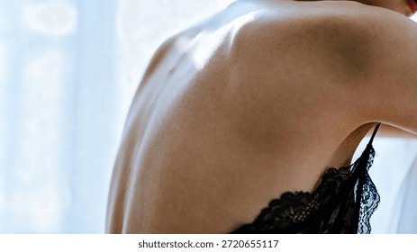 This image features a close-up view of a woman's shoulder and upper back, clad in a delicate black lace top. Soft, natural light highlights her skin, creating a warm and intimate atmosphere. The focus