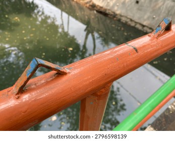 This image features a close-up view of an orange railing with a rusty metal piece attached to it, situated by a body of water. The railing is made of metal and has a glossy finish. In the background, a green body of water with fallen leaves and a concrete wall is visible. The overall atmosphere suggests an outdoor setting, possibly a park or a waterfront area.